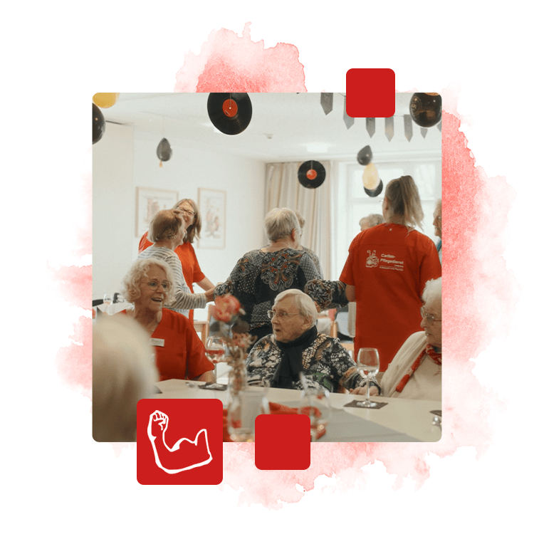 Foto von Senioren die an einem Tisch sitzen und sich unterhalten und lachen. Im Hintergrund stehen Senioren und Mitarbeiter Hand in Hand.